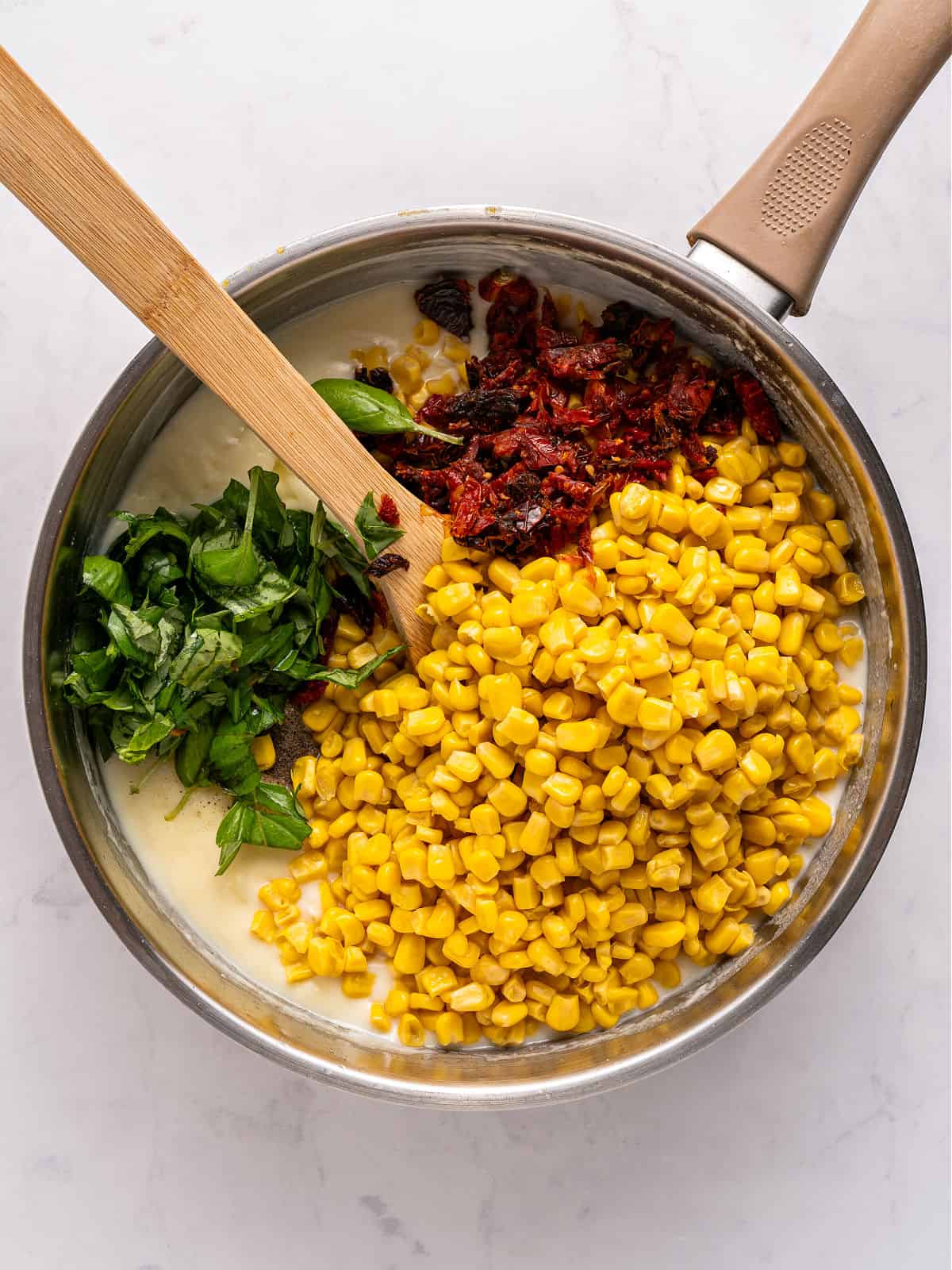 Basil, dried tomatoes and corn added to white sauce in metal pot.