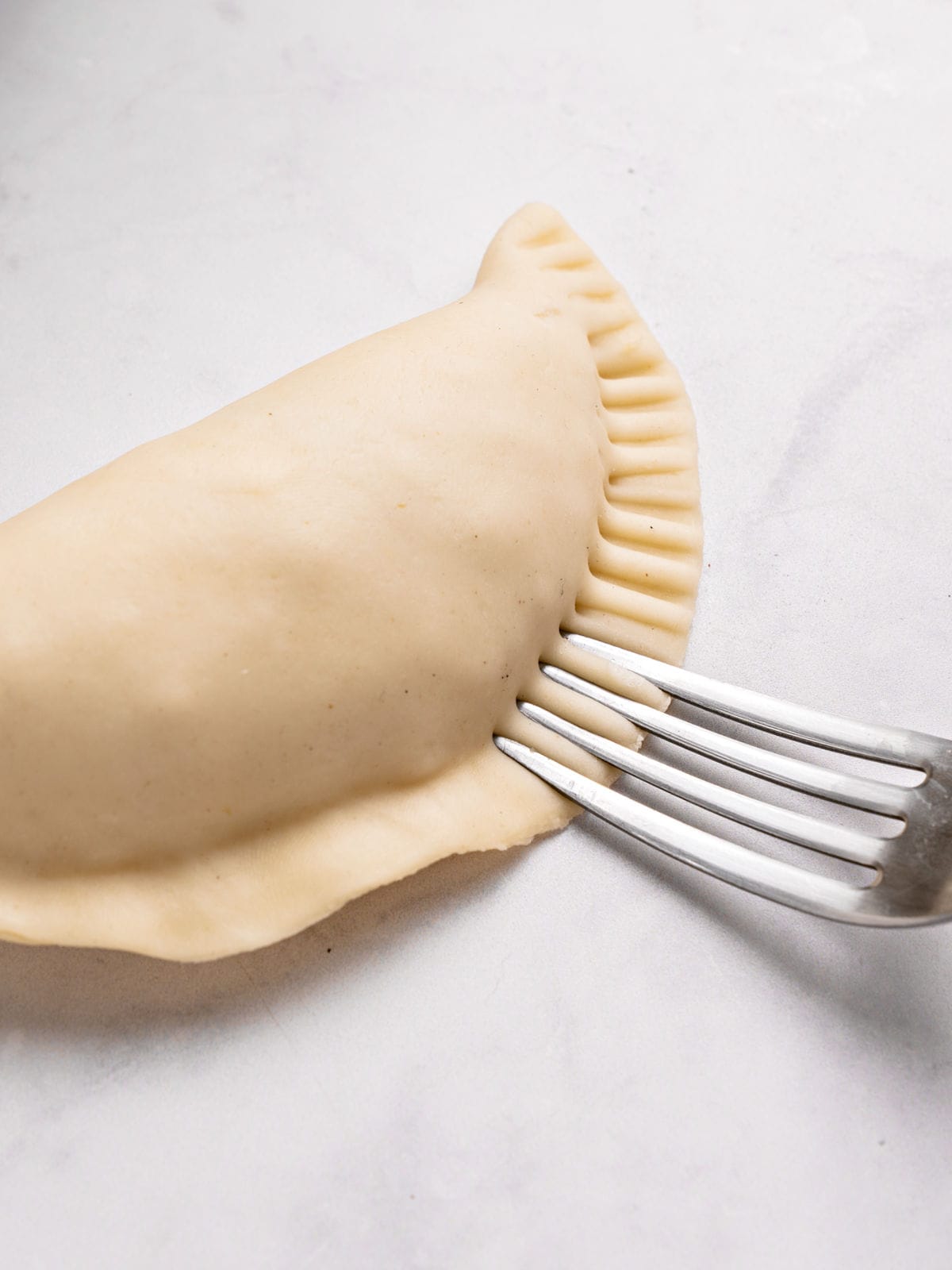 Crimping an empanada with a silver fork. White sauce with onion in a metal saucepan. White background.