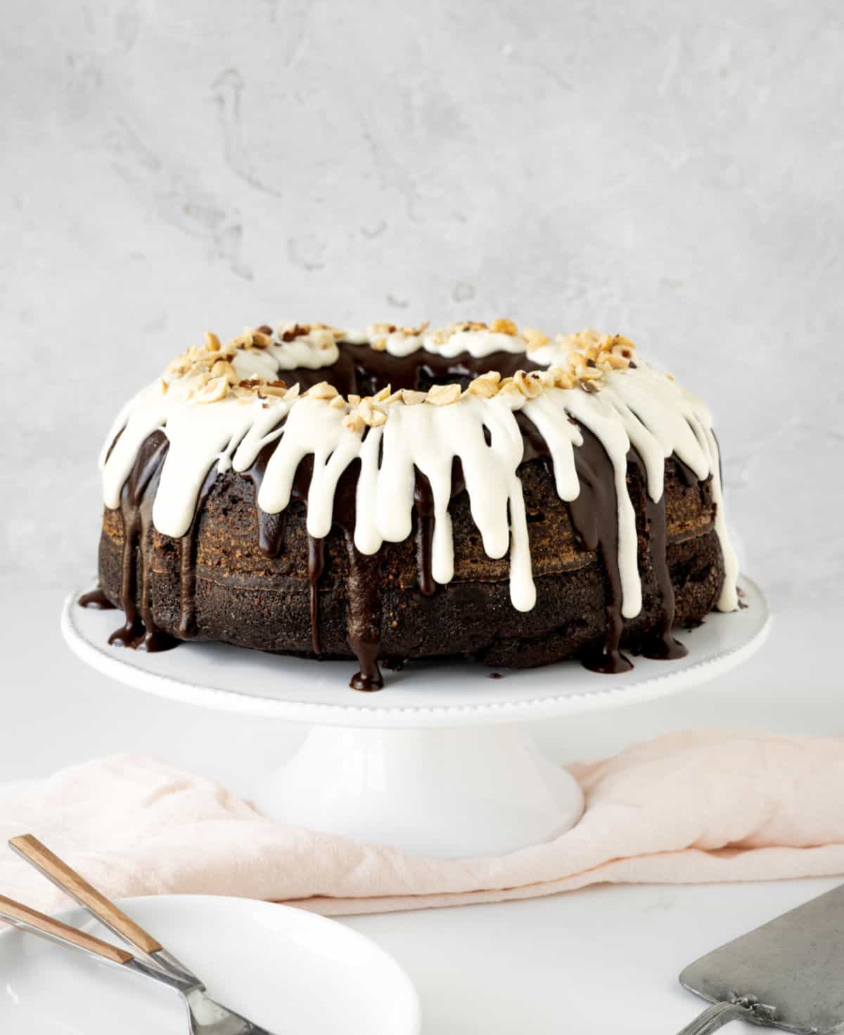 Whole dark and white glazed chocolate bundt cake on a white cake stand. Grey background, light pink cloth, plates, cake server. 