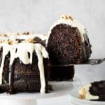 Double glazed chocolate bundt cake on a white cake stand with slice being lifted on a cake server. Grey background.