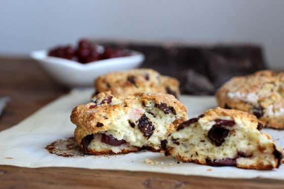 Halved chocolate sour cherry scones on white paper, bowl with cherries in background. 