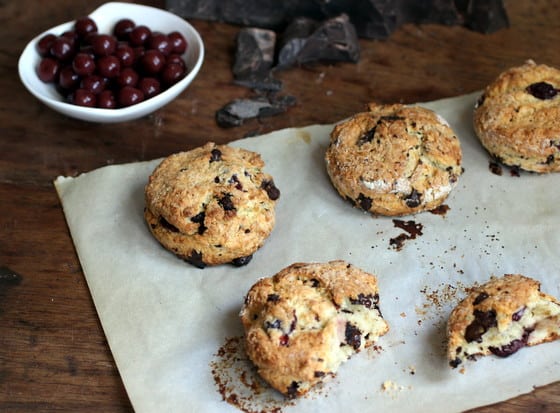 Chocolate sour cherry scones, whole and halved on parchment paper on a wooden table. A bowl of cherries. 
