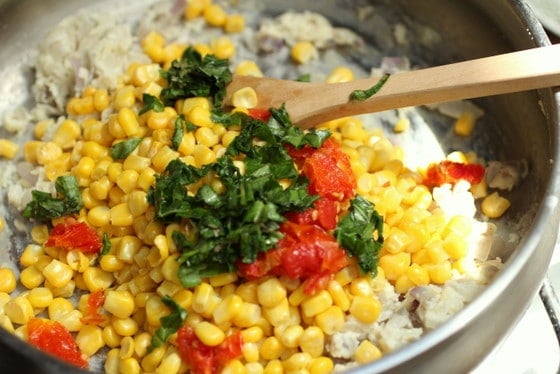 Basil, corn, and tomatoes added to white sauce in skillet. A wooden spoon.