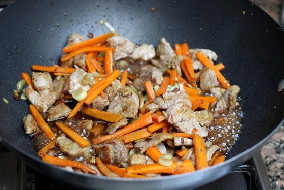 Pork bites with carrot stick and sauce being cooked in a dark metal wok.