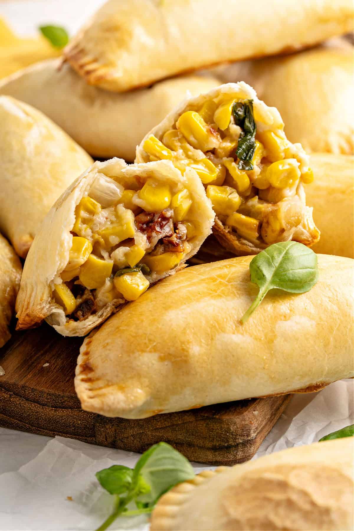 Two corn empanada halves on whole ones. Basil leaves. Wooden board.