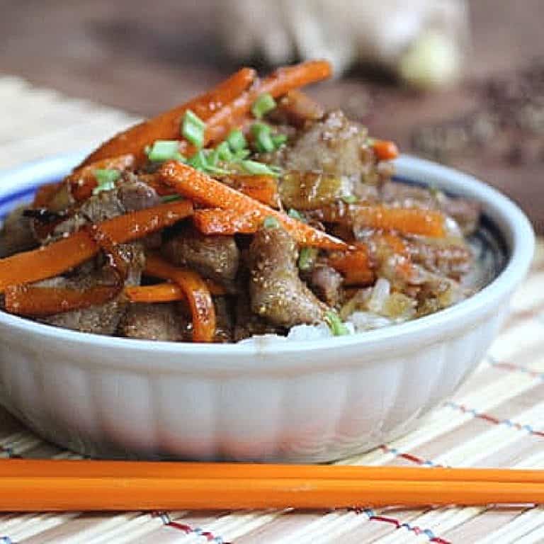 Close up of white bowl with pork and carrot stir fry on bamboo place mat.