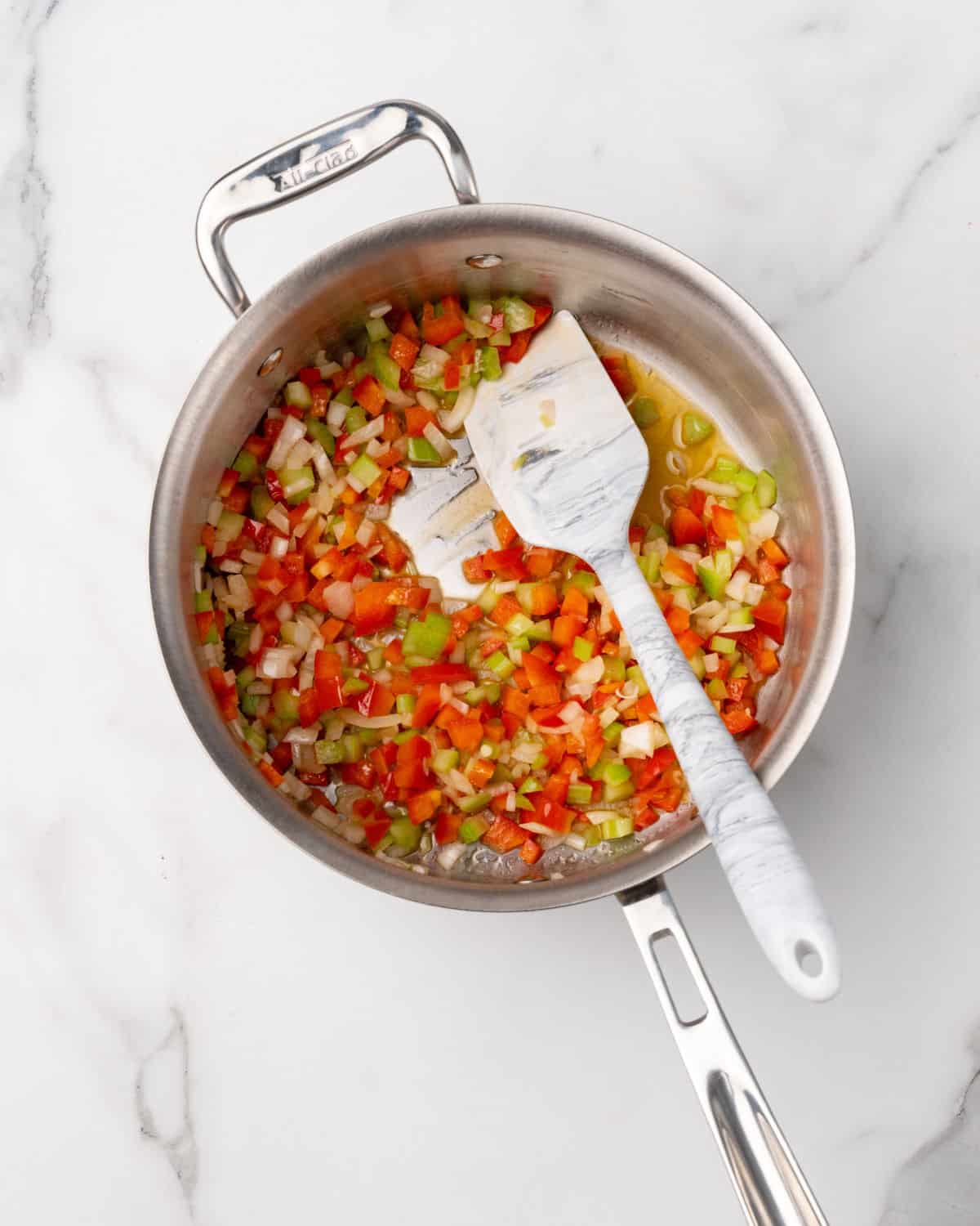 Metal pot with diced vegetables and a spatula on a white marble surface.