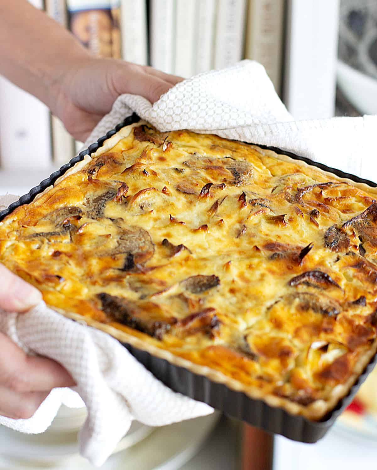 Hands holding square mushroom quiche still in a metal pan.