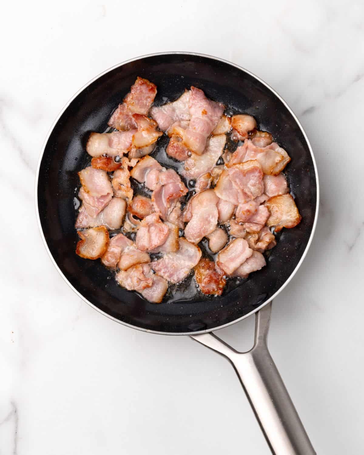 Bacon strips rendering on a black and metal skillet. White marble surface.
