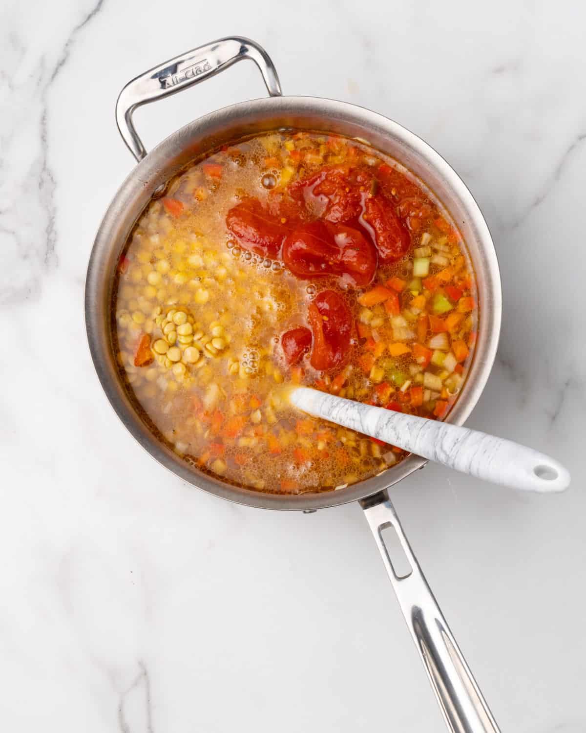 Tomatoes, diced vegetables, lentils, and liquid in a large metal pot with a white spatula inside. White marble surface. 