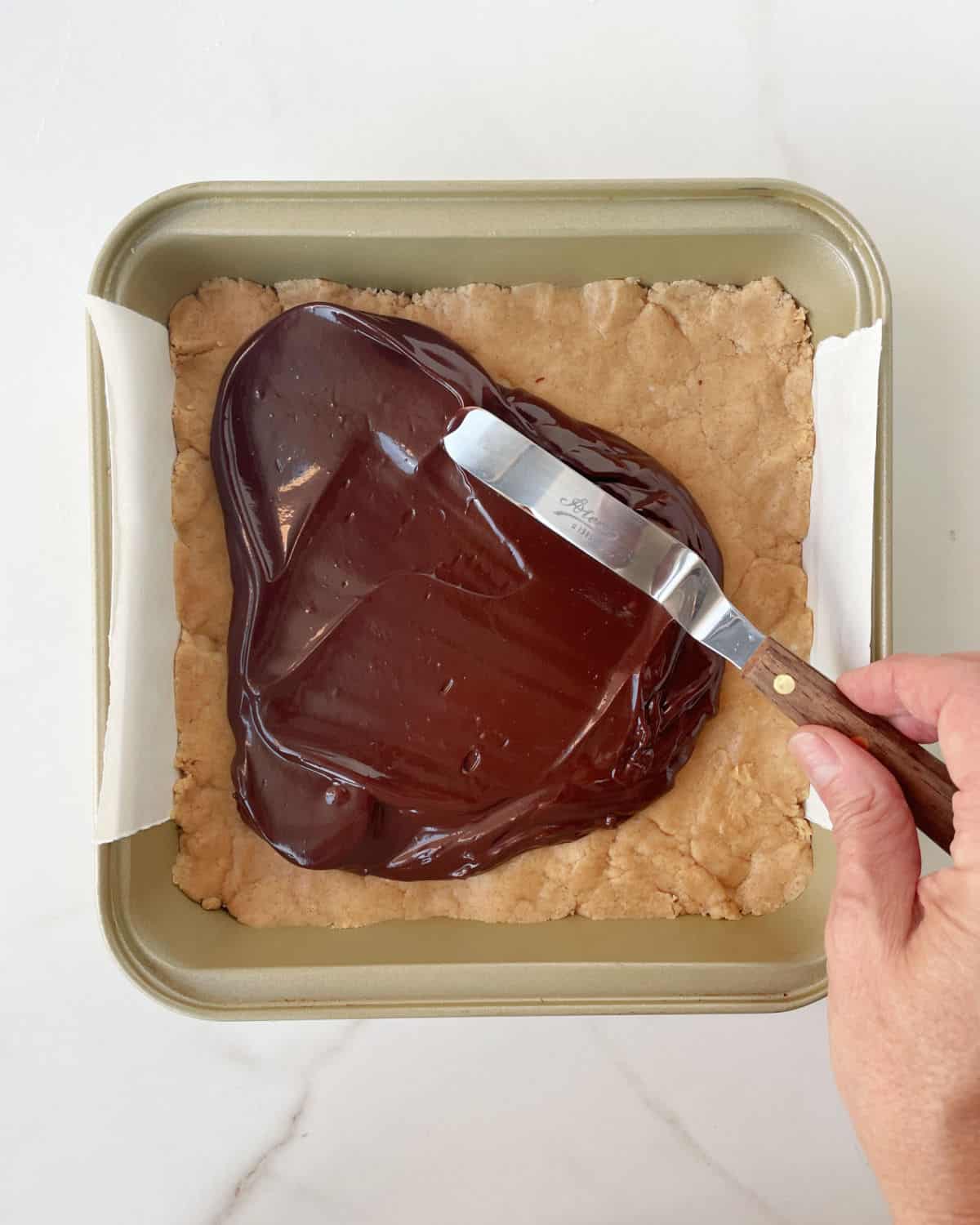 Spreading chocolate ganache with a spatula over peanut butter fudge layer in a square pan. White surface.