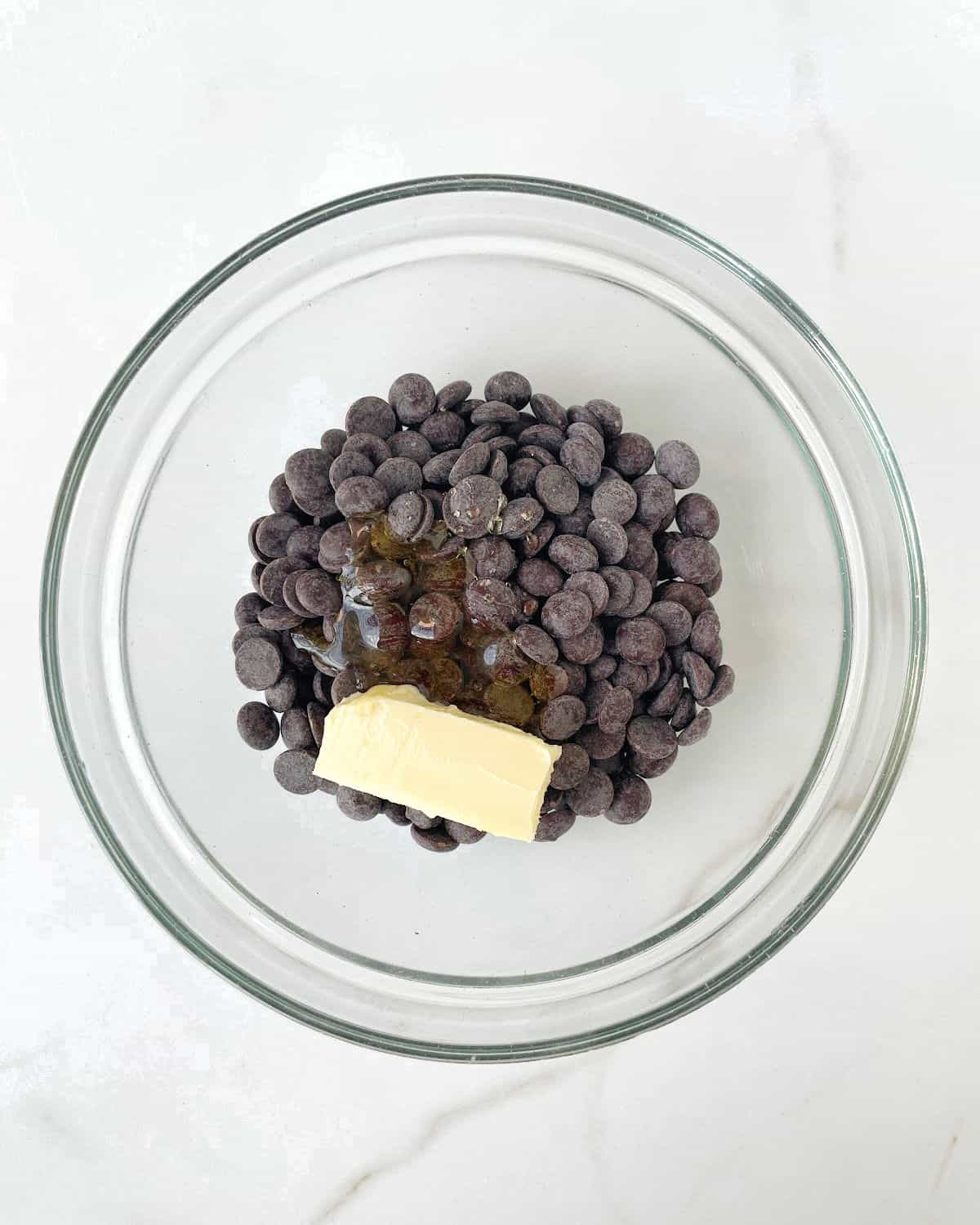 Chocolate chips and chunk of butter in a glass bowl on a white marble surface.