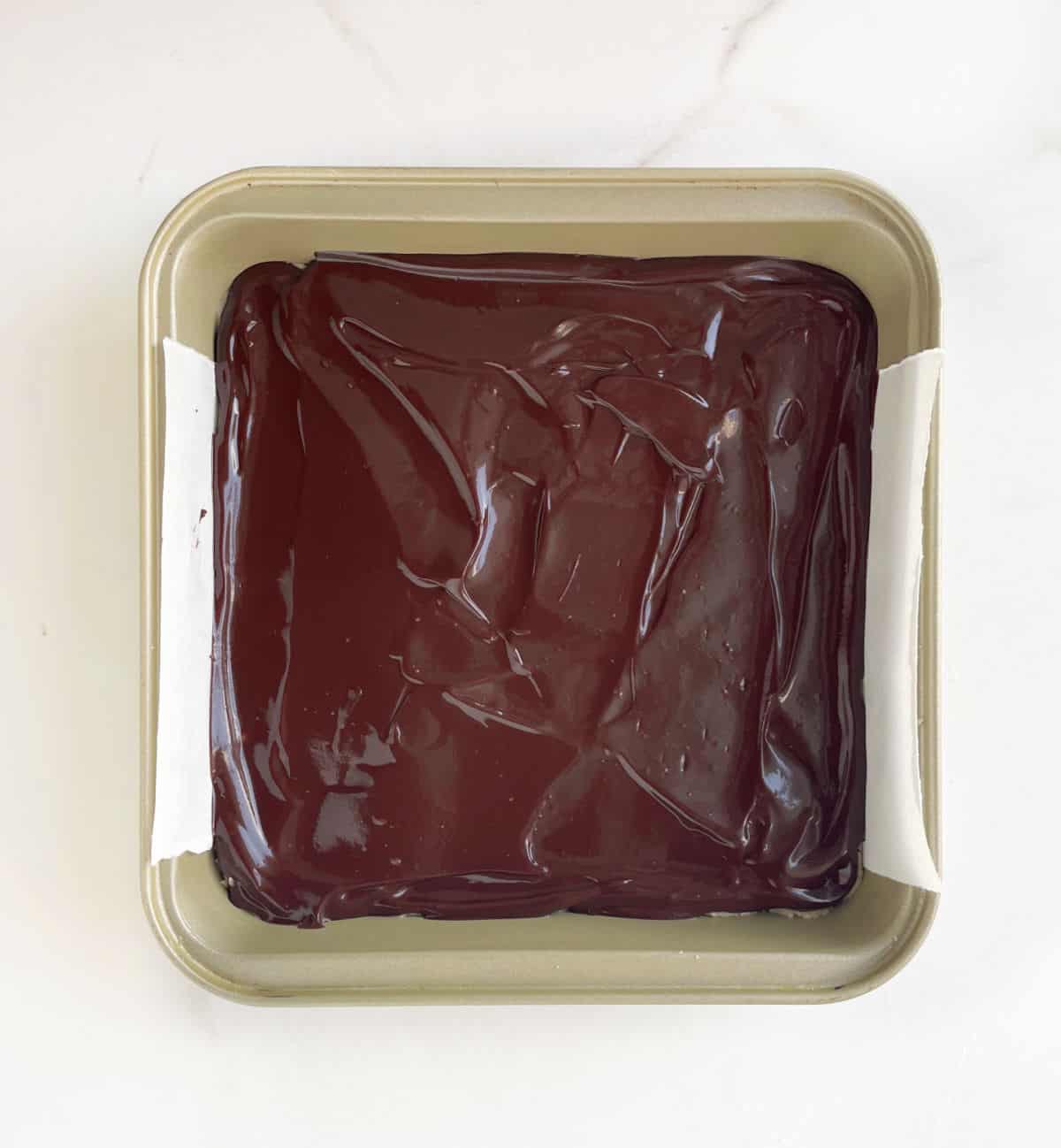 Chocolate coated fudge in a square baking pan on white marble.
