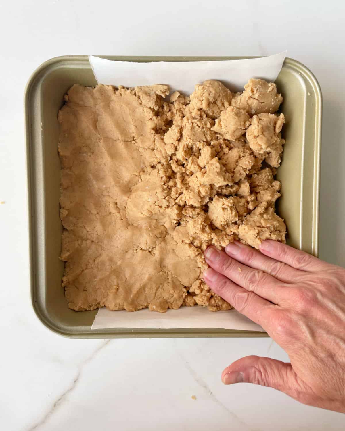 Hand patting peanut butter fudge layer on a golden square pan on a white marble surface.