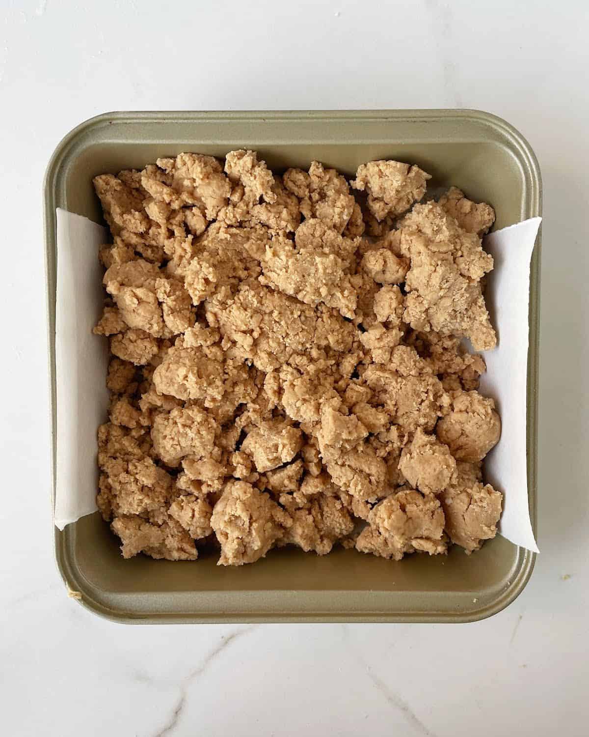 Crumbly peanut butter mixture in a gold colored square pan with parchment paper on a white surface.