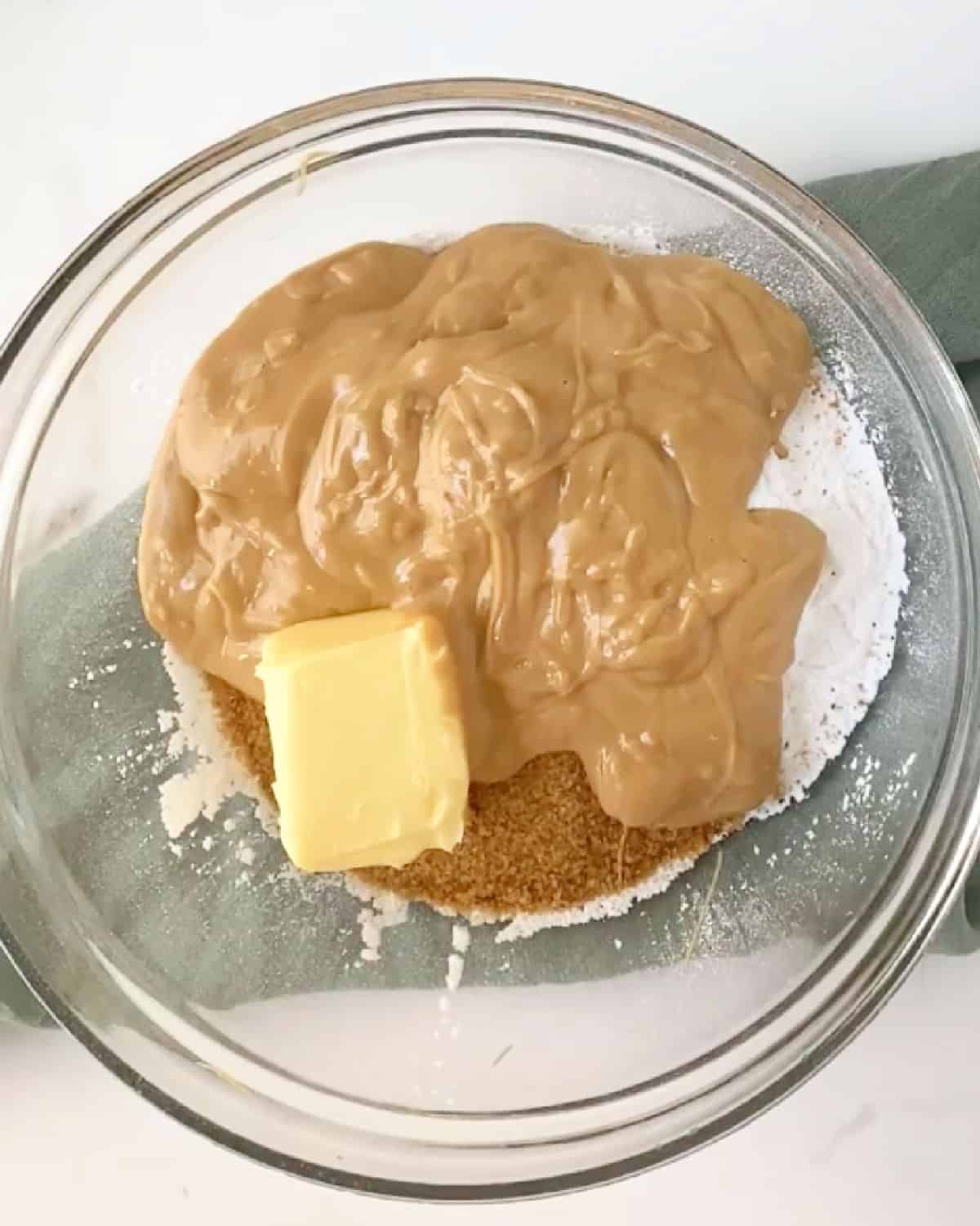 Peanut butter, butter, and powdered sugar in a glass bowl on a green cloth and white surface. 