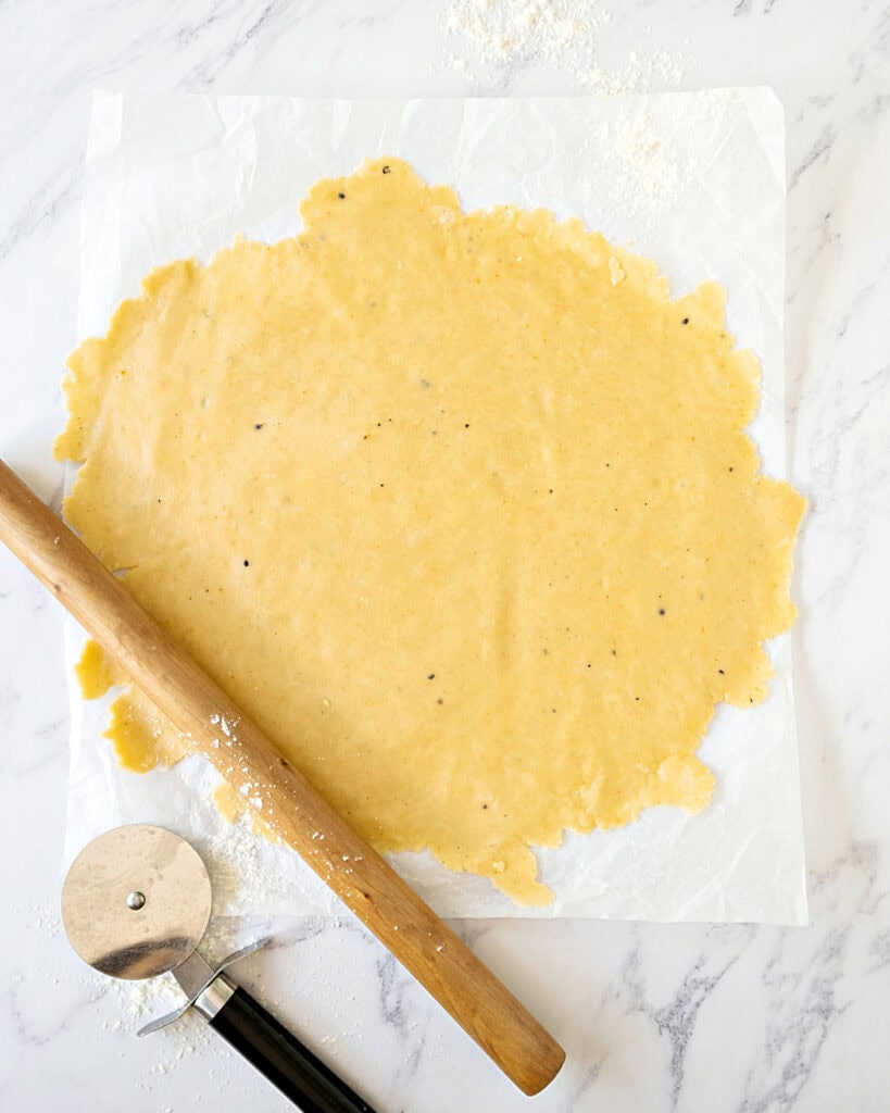 Rolled cheese cracker dough on a white paper and surface. Rolling pin and pizza wheel. 