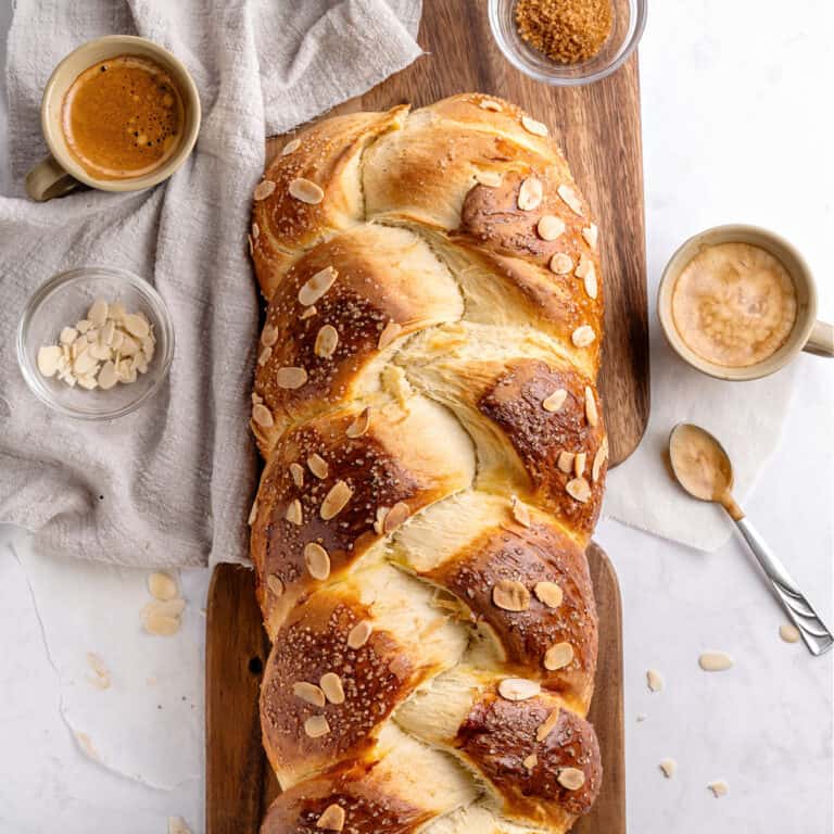 Partial top view of braided bread with almonds on a dark board. Light gray cloth, brown sugar bowl.