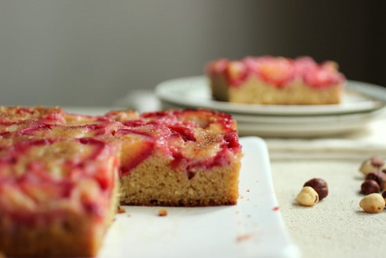 Partial view of plum cake on white plate with serving in stack of plates in the background.