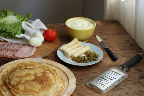 Spread of ingredients for savory crepe cake on wooden table, including ham, eggs, lettuce, mayo, tomato and more.