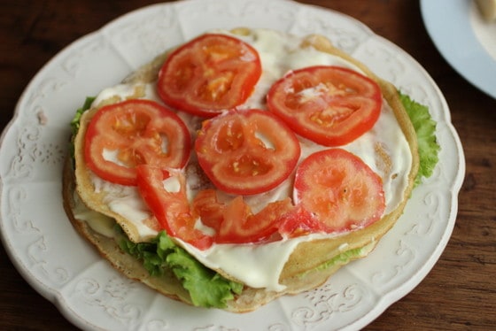 Building crepe cake with slices of tomato, mayonnaise and lettuce on white plate.