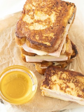 Top view of stack of ham and cheese French toast on beige parchment paper. A bowl with syrup.