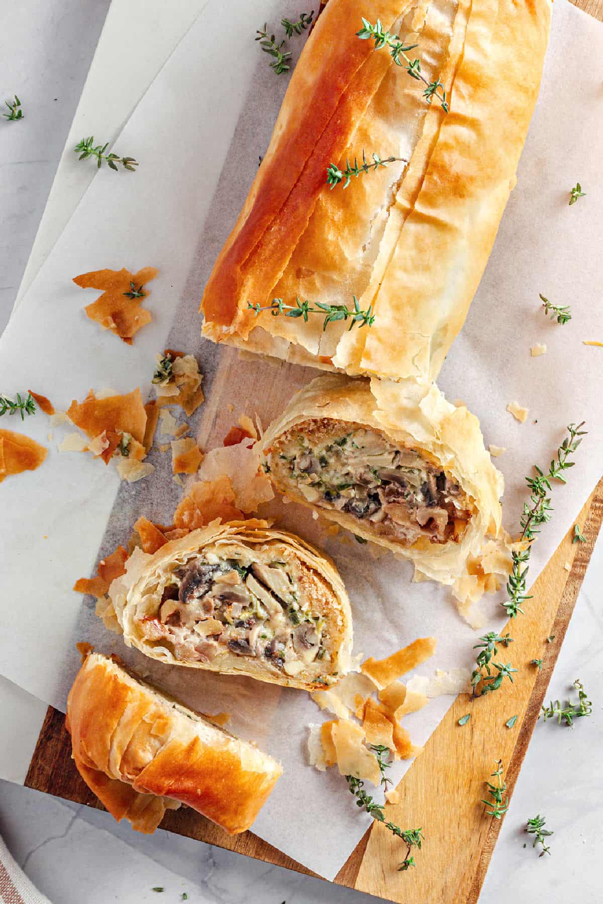 Mushroom Cheese Strudel Top view of sliced and whole mushroom phyllo strudel on wooden board with parchment. Thyme sprigs.