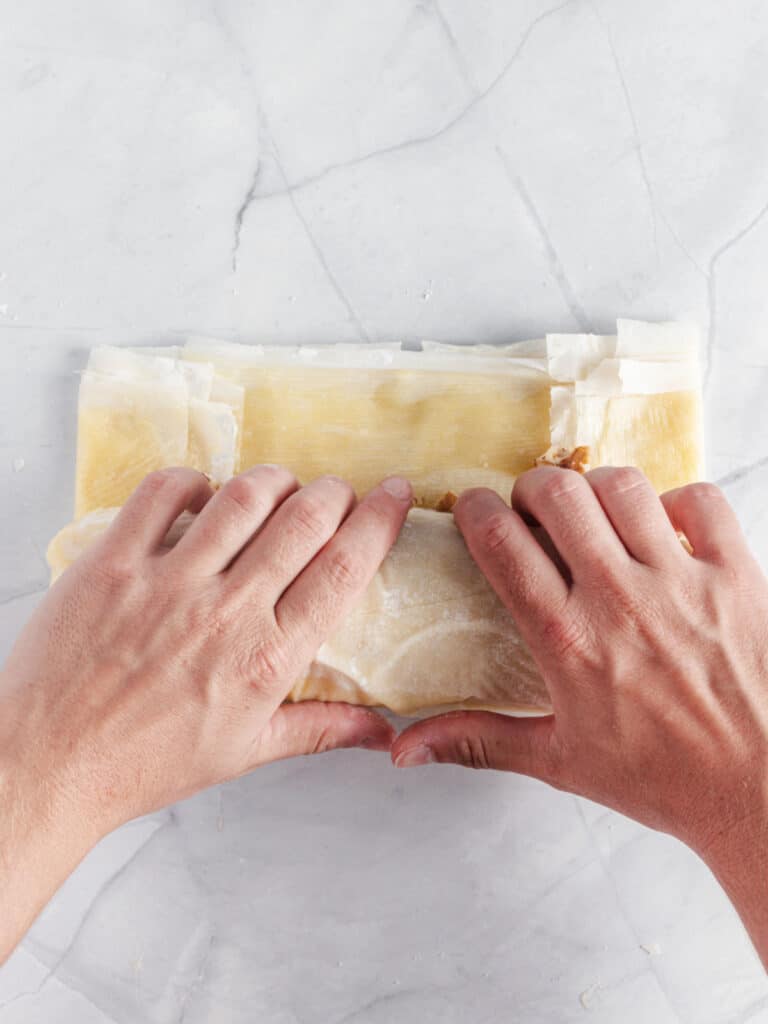 Hands rolling phyllo dough strudel on a white marbled surface.