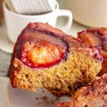 Close up slice of plum upside-down cake being lifted. Cups in the background.
