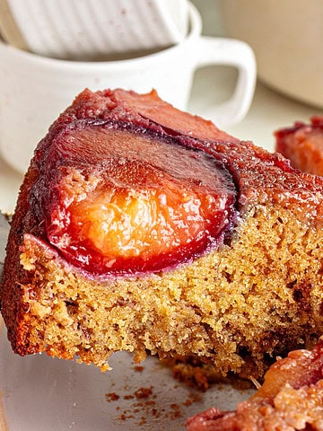 Close up slice of plum upside-down cake being lifted. Cups in the background.