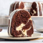 Close up slice of cheesecake stuffed chocolate bundt cake on a plate. Whole cake in the gray background.