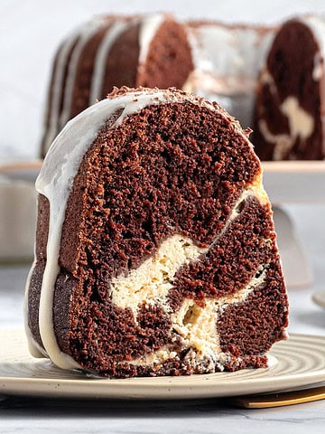 Close up slice of cheesecake stuffed chocolate bundt cake on a plate. Whole cake in the gray background.