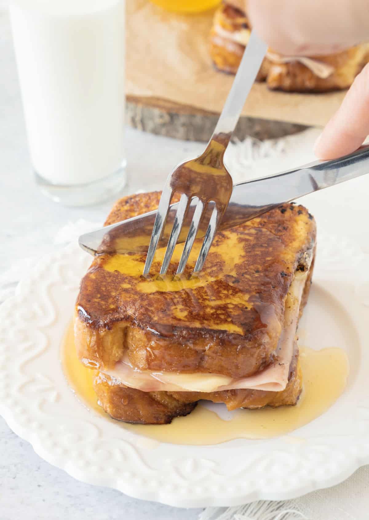 Cutting a syrup drenched ham and cheese French toast sandwich on a white plate. 