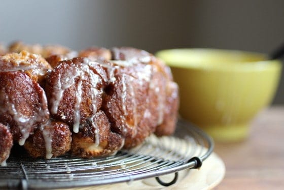Glazed monkey bread on a wire rack on a wooden surface, with yellow bowl in background. 