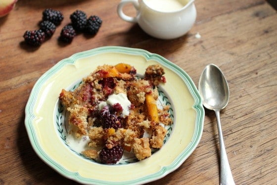 Peach Brown Betty serving on yellow green plate on wooden board. A spoon, cream jar, and blackberries. 