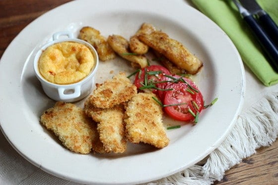 White plate with chicken tenders, banana, corn pudding and tomato slices