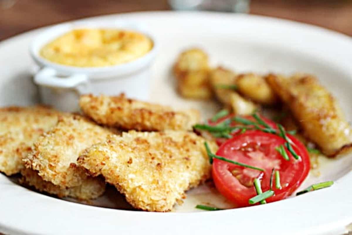 Chicken tenders, tomato slices, ramekin with corn pudding and banana on a beige plate.