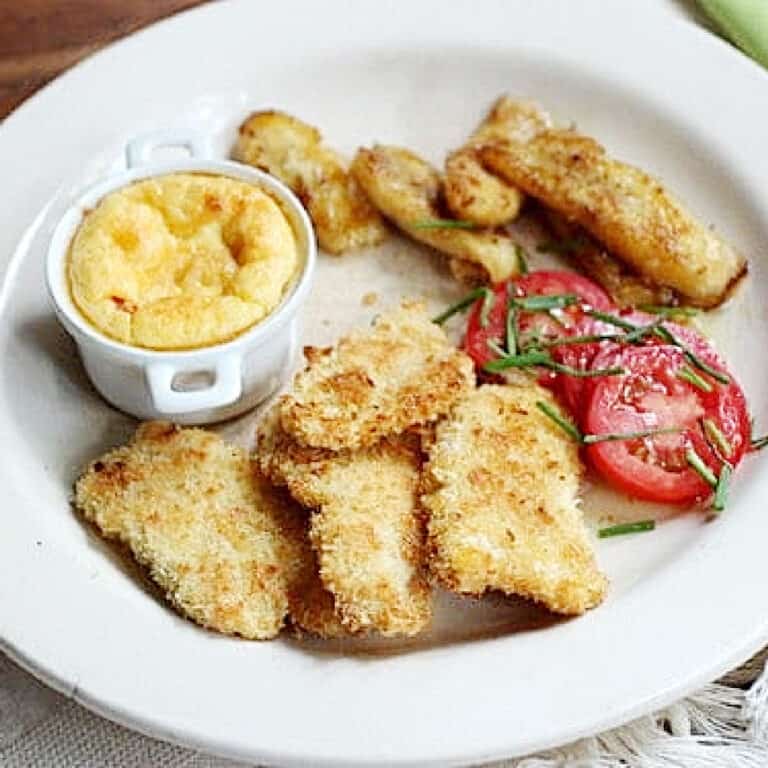 Beige plate with chicken tenders, corn pudding, fried banana pieces and sliced tomatoes.