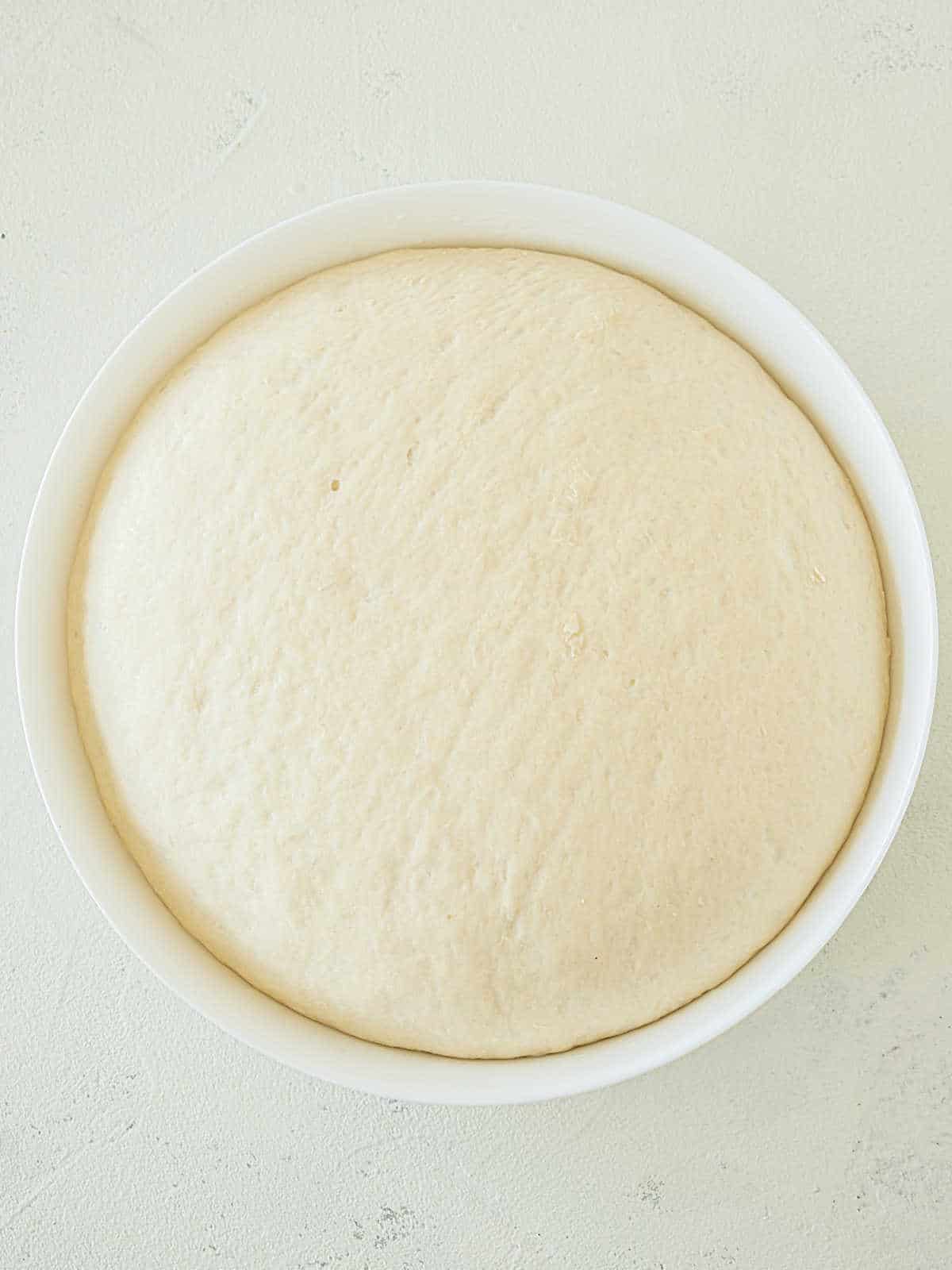Proofed pretzel dough in a white bowl on a whitish surface.