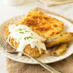 White plate with breaded chicken cutlet, white gravy, corn pudding and bananas. Brown and white surface.