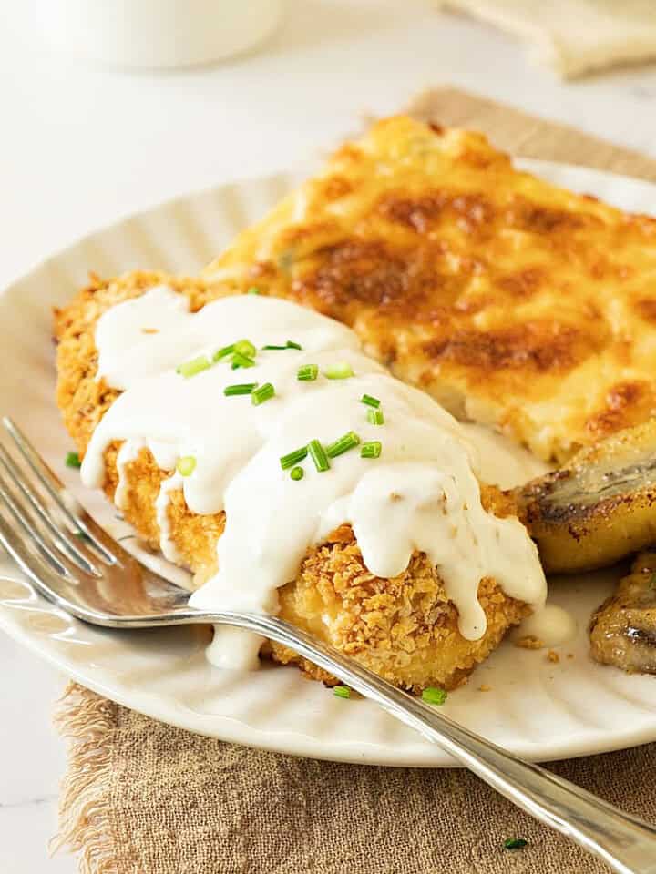 White plate with breaded chicken cutlet, white gravy, corn pudding and bananas. Brown and white surface.