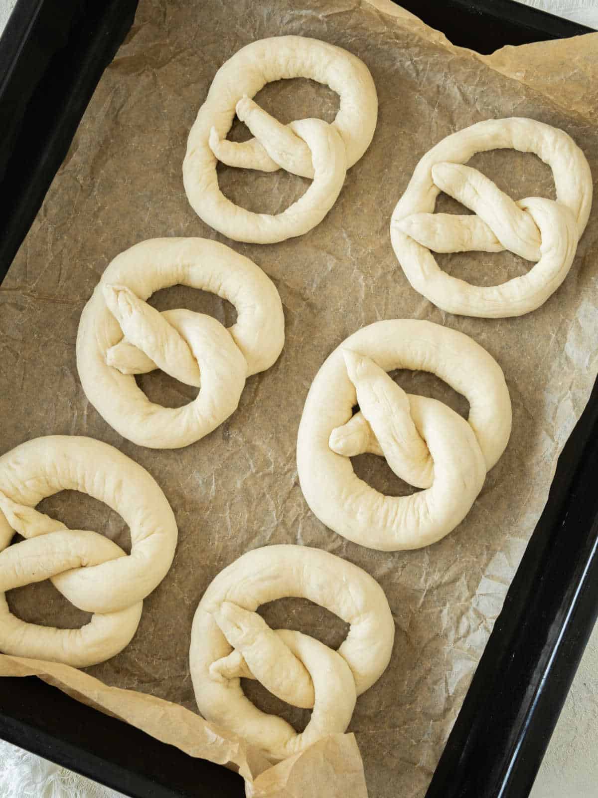 Formed pretzels on beige paper on a black oven tray.