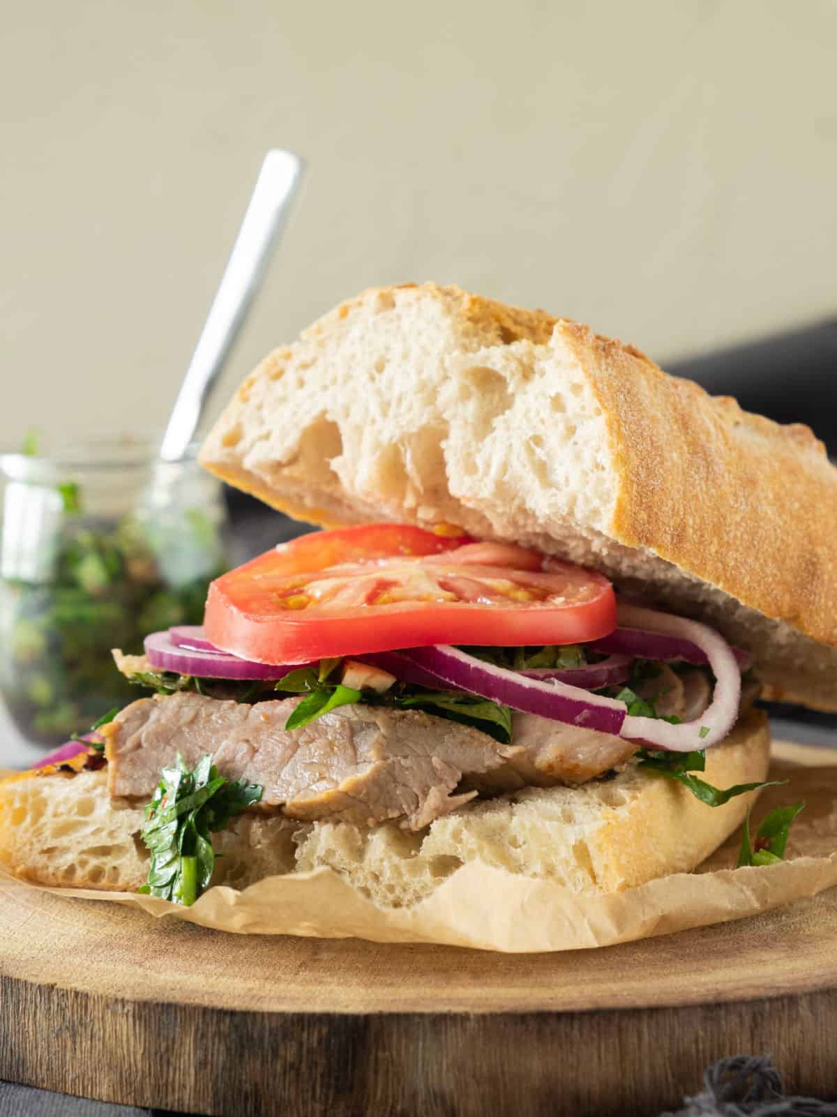 Steak sandwich with tomato and onion on a wooden board. A chimichurri jar in the background.