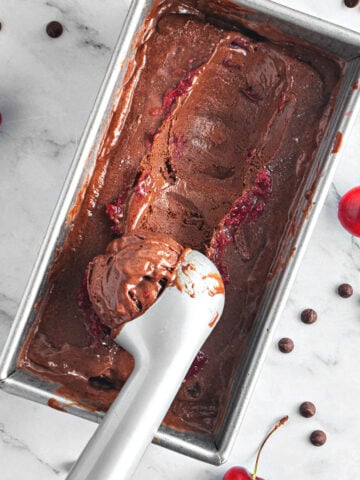 Scooping chocolate ice cream with cherries in a metal loaf. White marbled surface, loose chips, cherries.