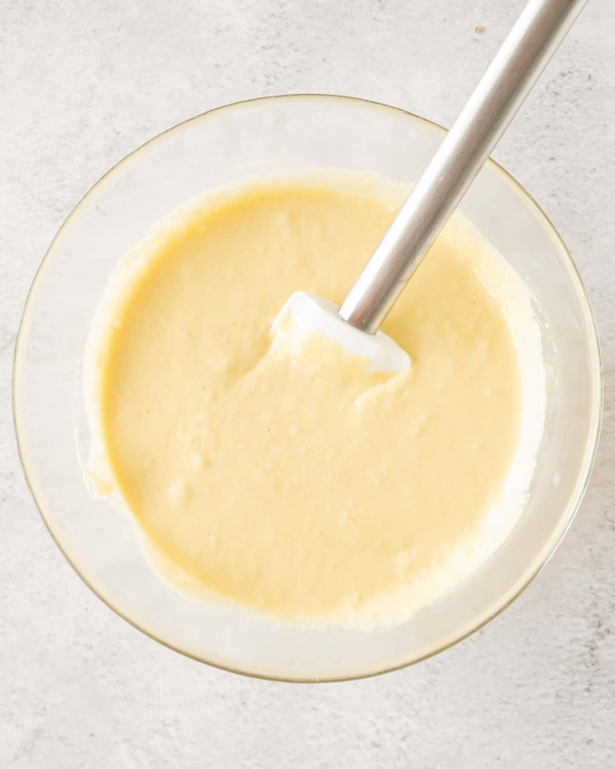 Glass bowl with cornmeal pancake batter and silver spatula. Light grey surface. 