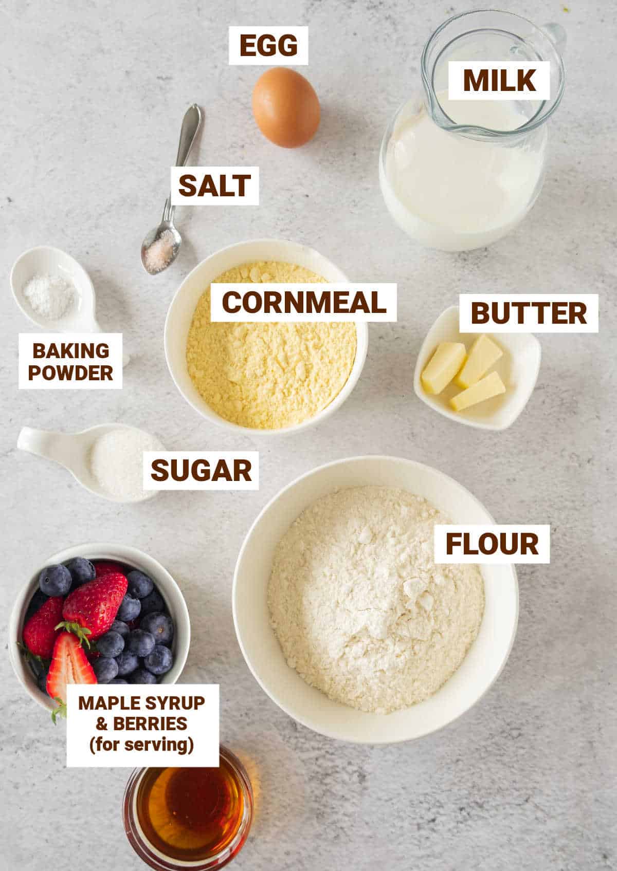 Grey surface with bowls containing ingredients for cornmeal pancakes including egg, milk, butter, syrup, berries, flour, salt.