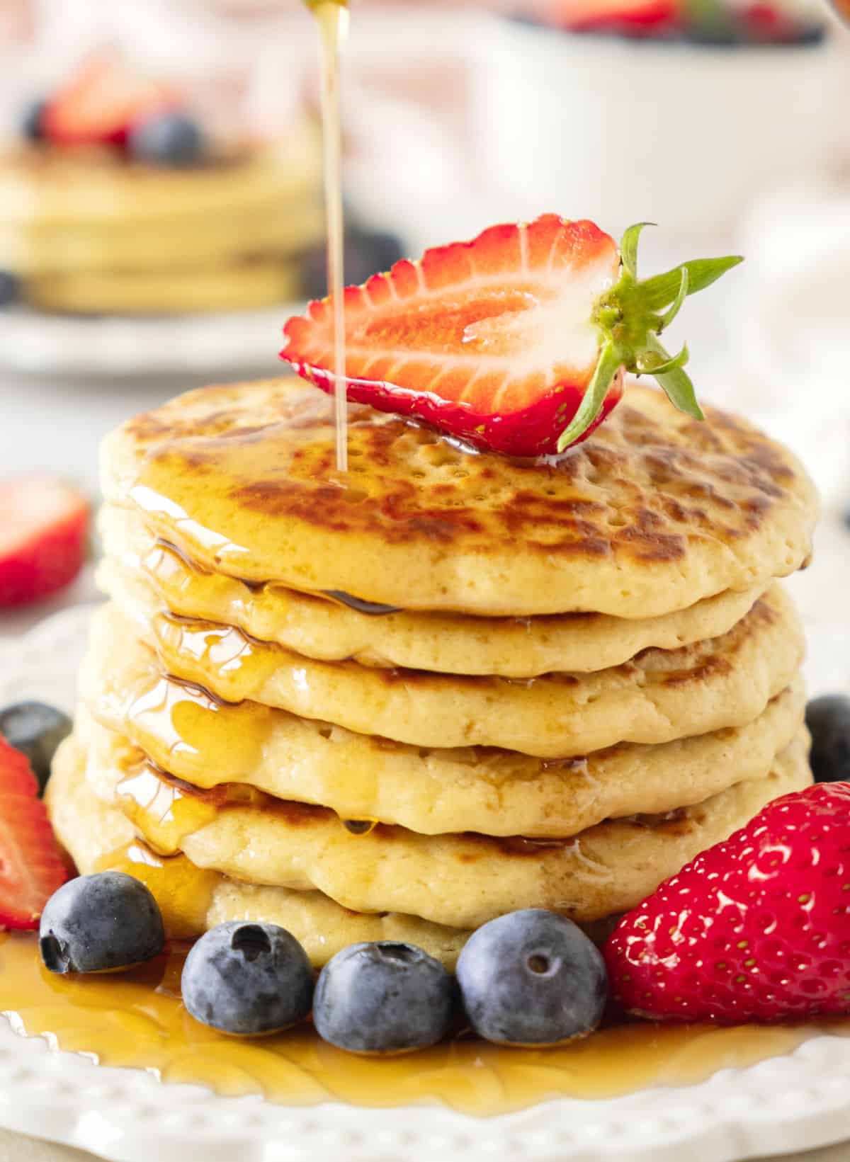Stack of pancakes in a white plate with fresh berries and syrup drizzling from the top.
