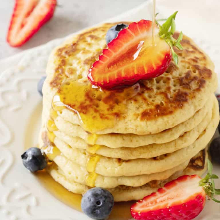 White plate with stack of cornmeal pancakes drizzling with syrup and fresh berries.