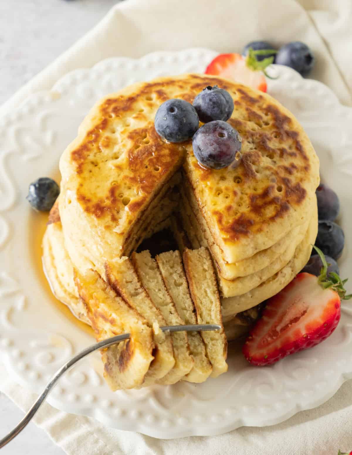 Several pancakes stacked in a white plate with syrup and berries, piece is cut and forked. 