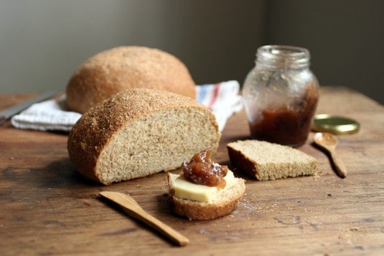 Loaves of whole wheat bread, slices with jam, jar, and utensils on a wood table. 